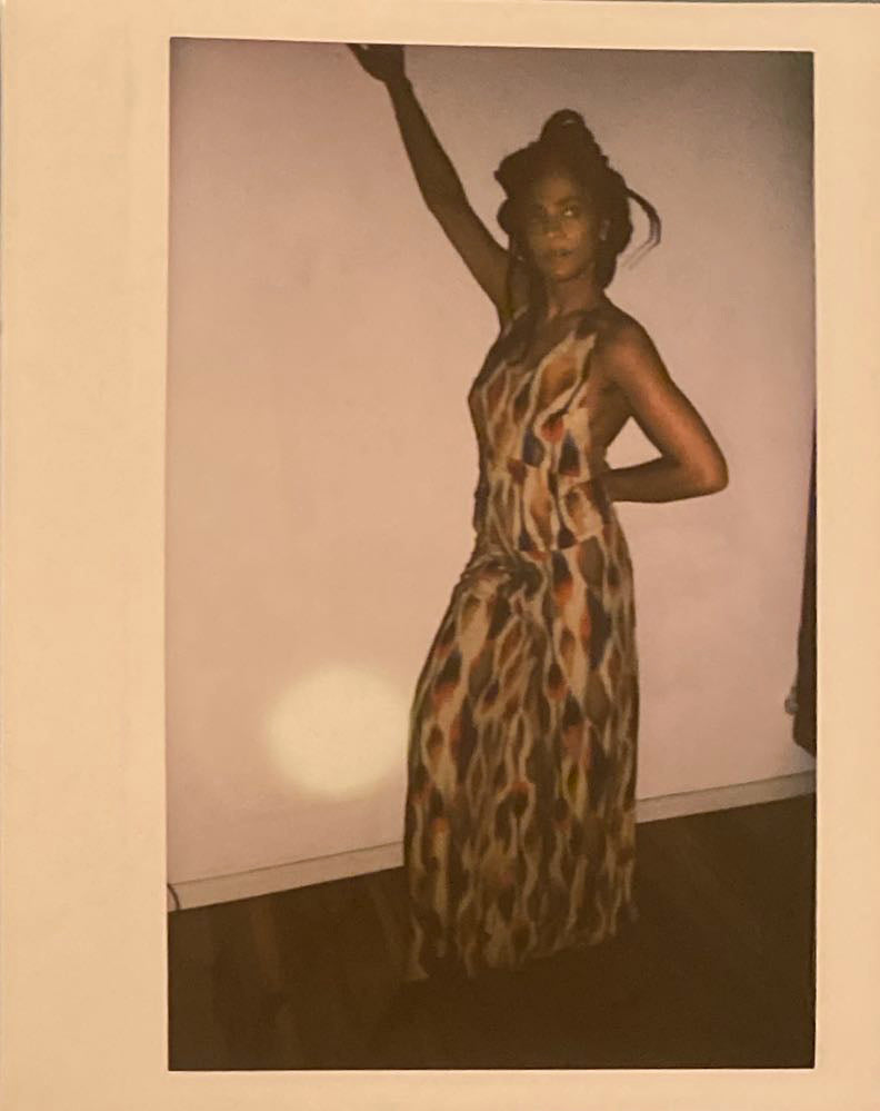 A polaroid of a woman standing against a wall with her hand up.