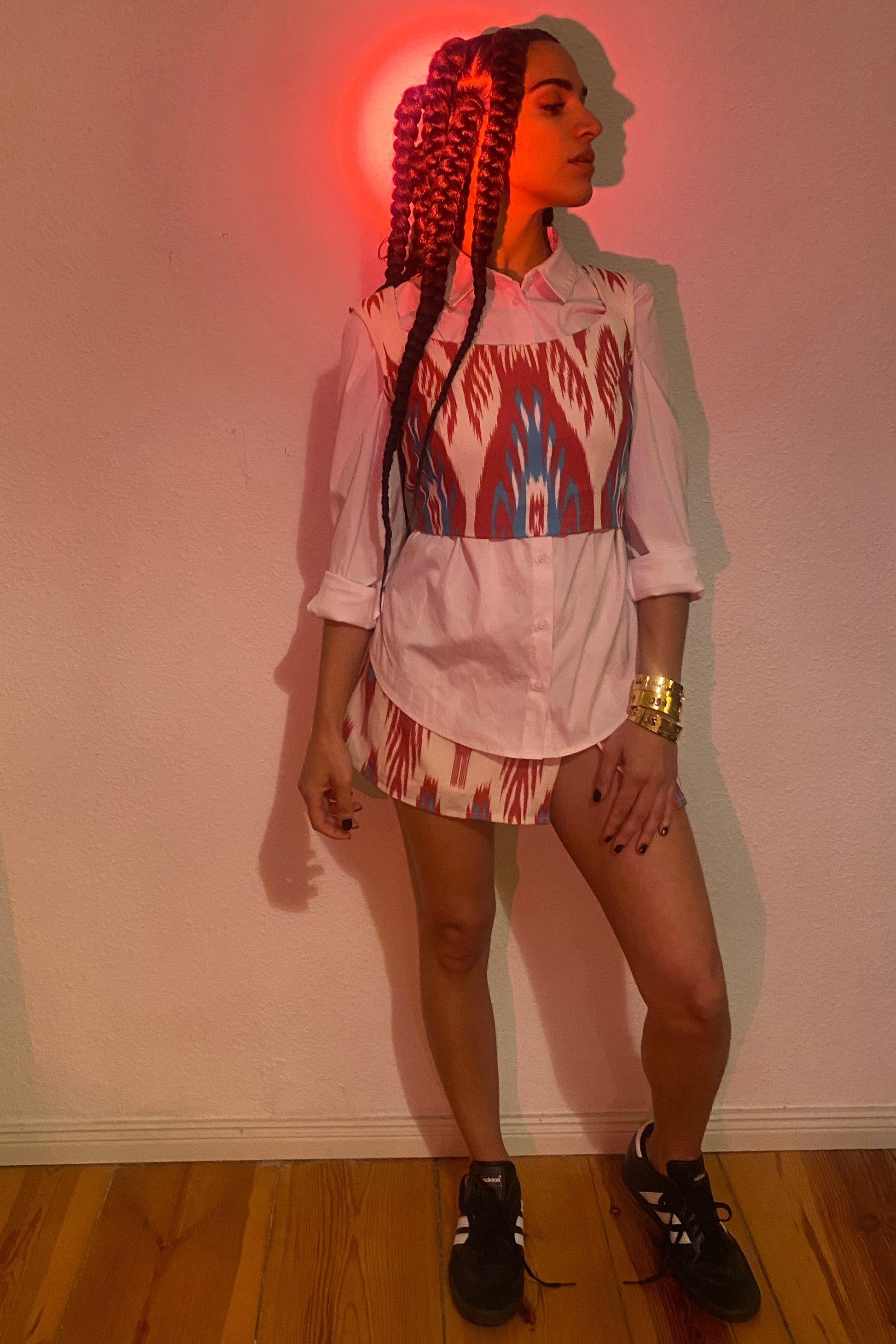 A red light shines on a woman with braids in a collared white shirt and ikat/adras mini skirt.