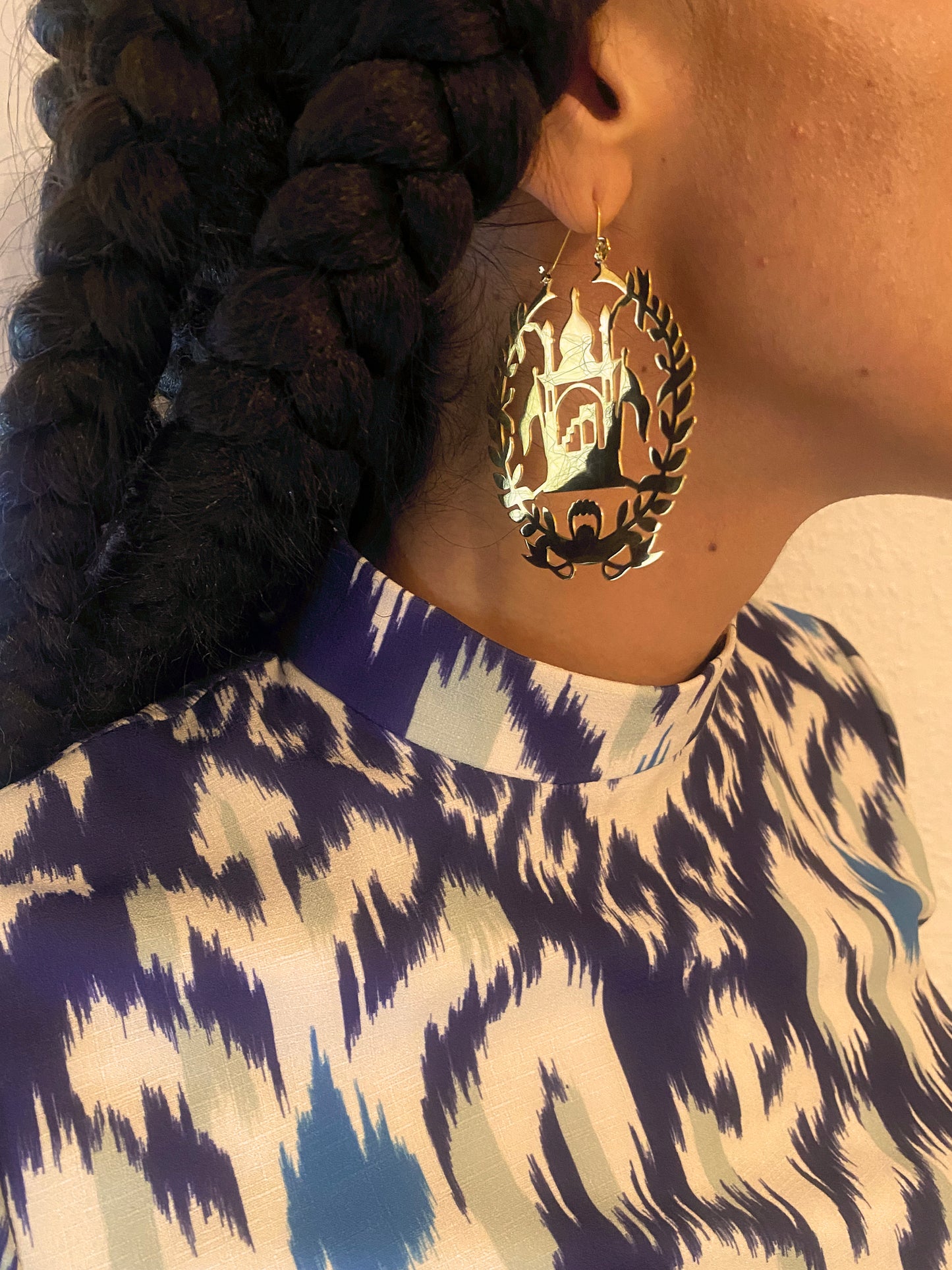 A woman with long braids wears gold national emblem shaped earrings reflecting light.